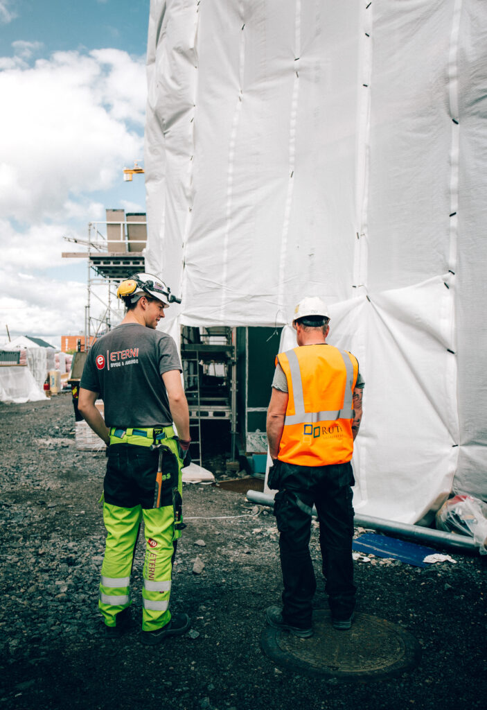 Bjørn Henrik står med ryggen til på en byggeplass i grønn arbeidsbukse, sammen med en kollega i oransje vest foran et stillas