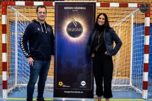 White man with dark brown hair stands in front of a handball goal, with a lady with dark hair, dressed in black on the right. Roll-up with Bergen Handball's logo in the middle.